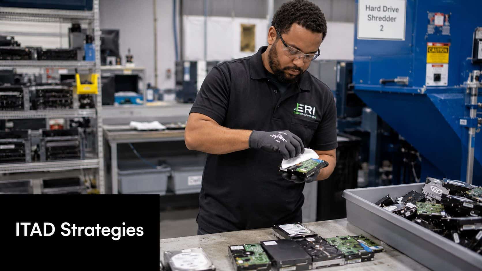 A technician wearing safety glasses and gloves cleaning a hard drive at an IT asset disposal facility.